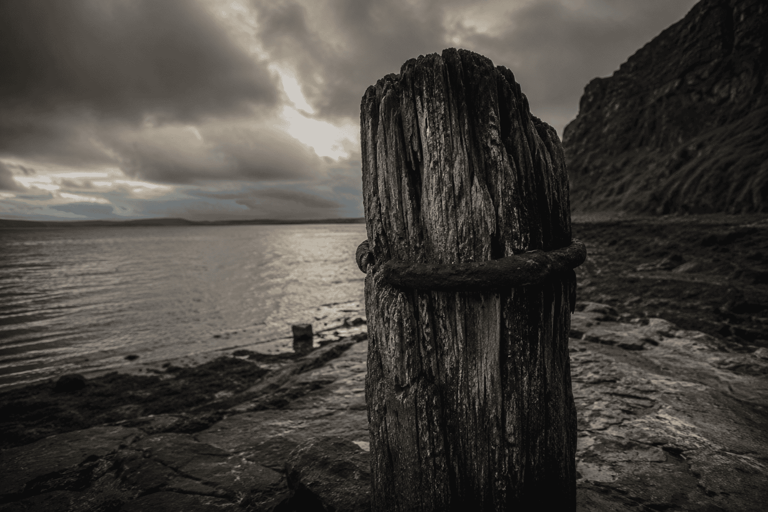 Old timber bollard with worn grain, fractured top, dark sky and lonely shoreline.